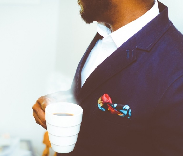 Groom in a tailored navy suit with white shirt and floral pocket square, holding a coffee