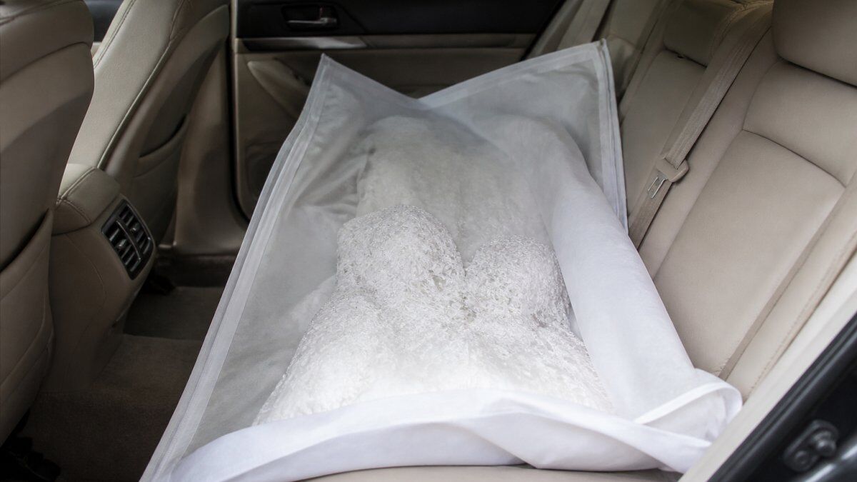 Wedding dress carefully laid flat in vehicle's back seat, protected by white tissue paper and layered in a clean garment bag, showing proper positioning and padding with natural car interior lighting