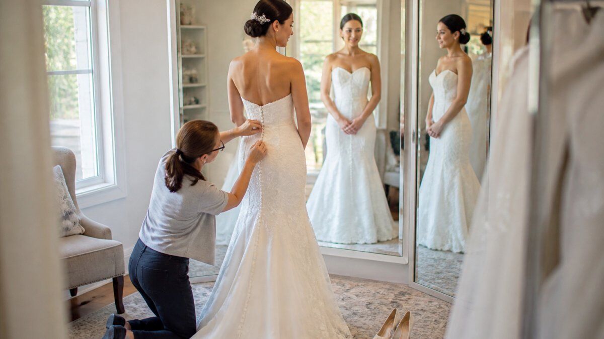 Bride in an elegant white wedding dress standing at a fitting appointment, a professional seamstress adjusting the bodice seams while the bride looks in a three-way mirror, with white wedding heels visible on the floor and natural studio lighting