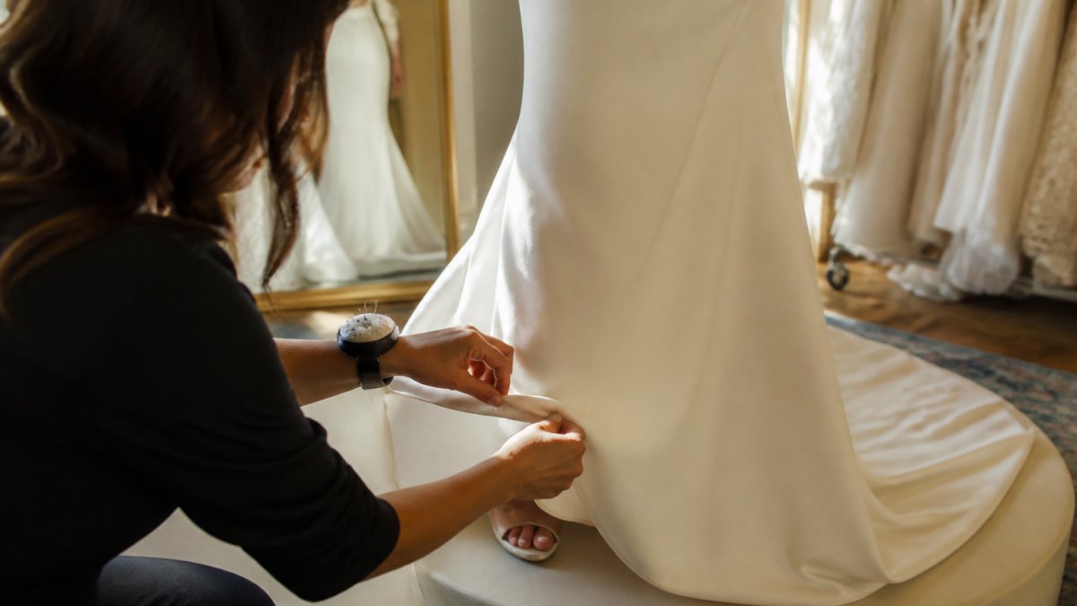 Seamstress kneeling to pin the hem of a bride's wedding dress on a pedestal in a bright studio